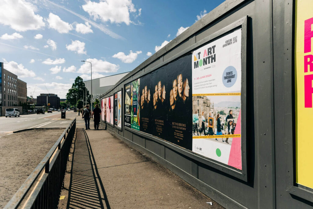 New Town Art Month posters displayed on a construction hoarding along a sidewalk.
