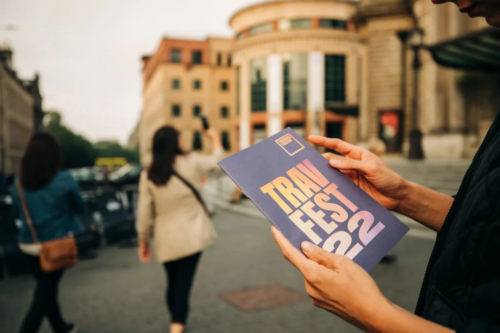 TravFest 2022 brochure held by a person with blurred people and buildings in background