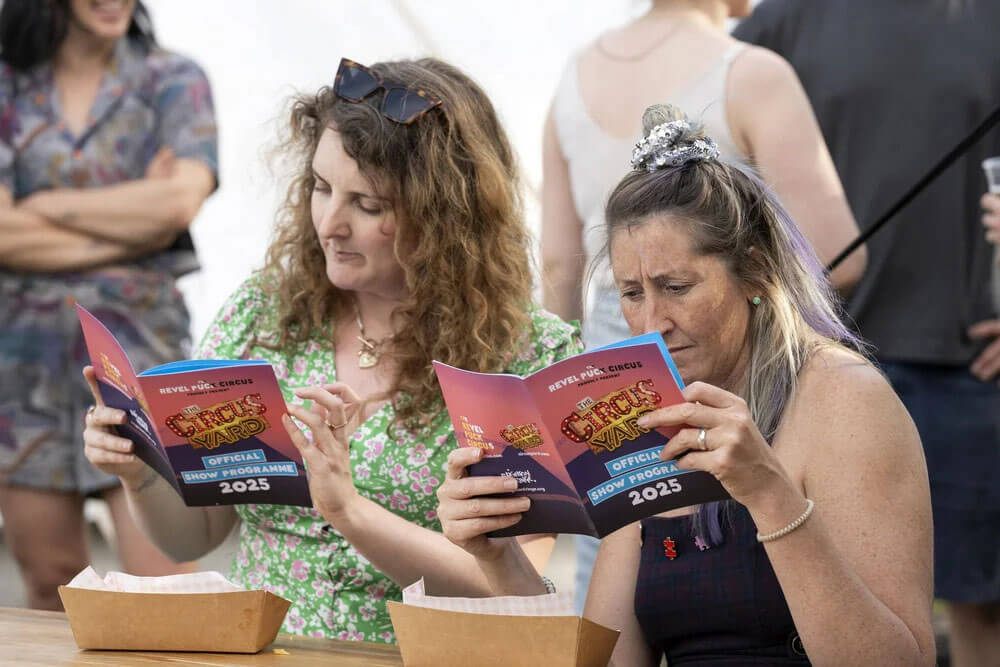 Two women read "The Circus Yard Official Show Programme 2025" at an outdoor event